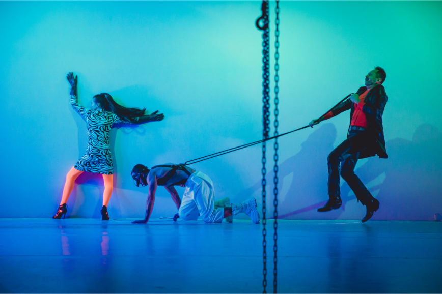 Niebieskie światło, trzy osoby. Z lewej strony kobieta odwrócona plecami, podpiera się o ścianę. Ma wzorzystą sukienkę i neonowo pomarańczowe rajstopy. Z prawej strony idzie mężczyzna w skórzanej kurtce i czerwonej koszuli. Na smyczy prowadzi klęczącego mężczyznę w białych spodniach.