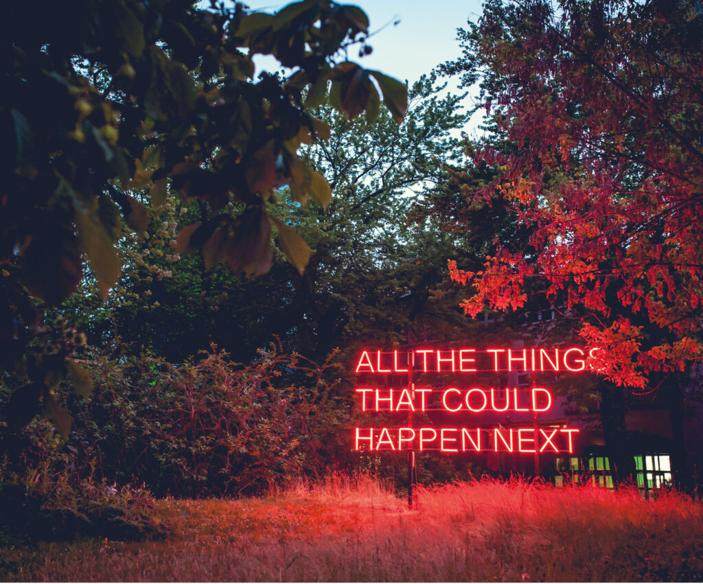 Czerwony neon z tekstem All the things that could happen next. Wokół niego drzewa i wysokie trawy oświetlone przez neon.