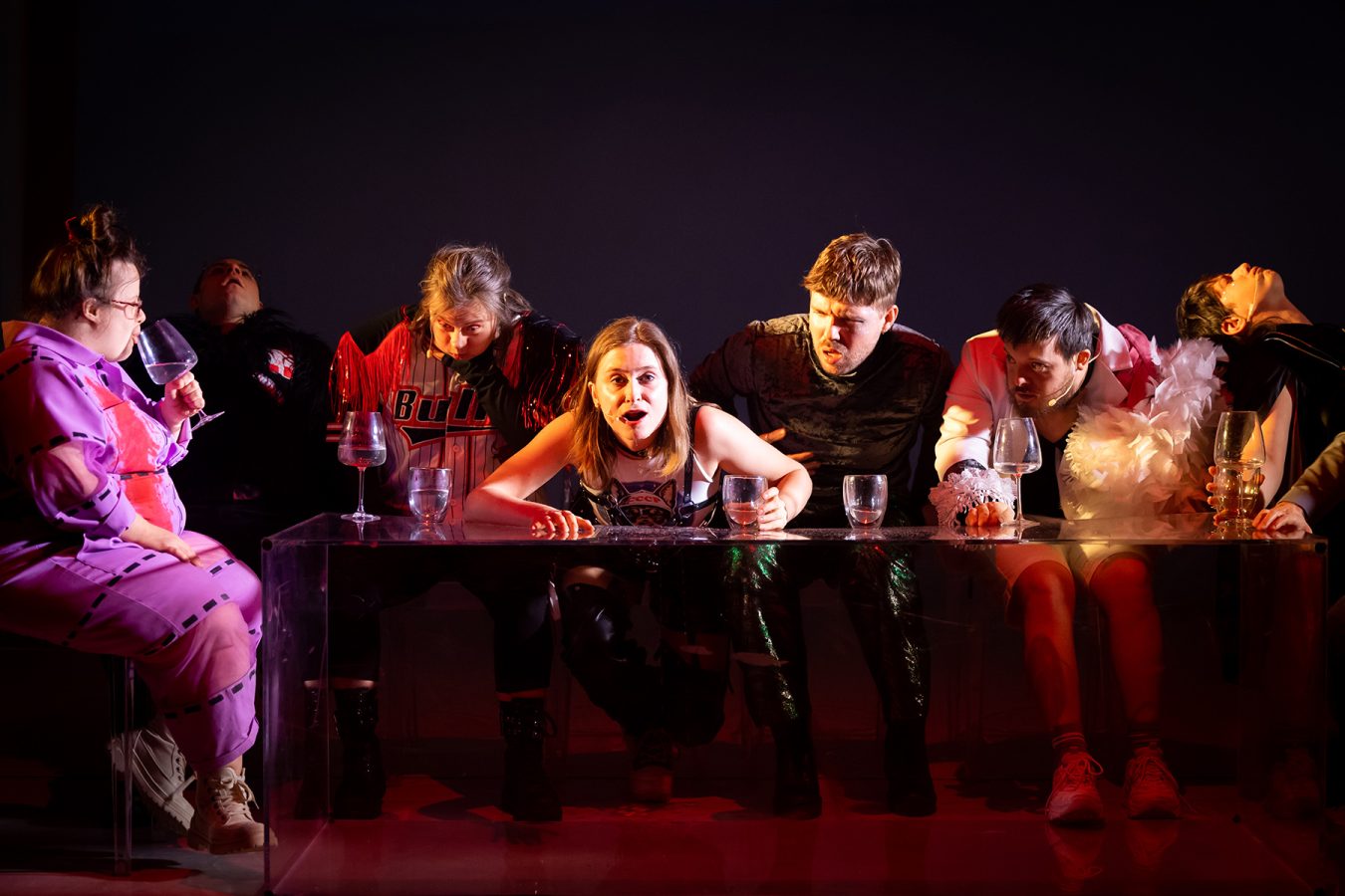 W kostiumach zwierzęcych aktorzy Teresa Foks, Piotr Sakowski, Aleksandra Skotarek, Helena Urbańska, Daniel Kotowski, Daniel Krajewski i Ania Nowak siedzą przy długim stole z przezroczystej pleksi - na stole stoją zdobne kieliszki