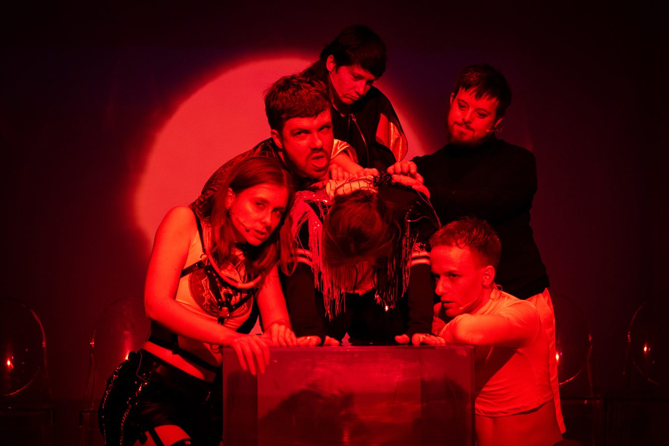 Pięcioro aktorów pochyla się na skuloną postacią –grupę oświetla czerwona plama światła